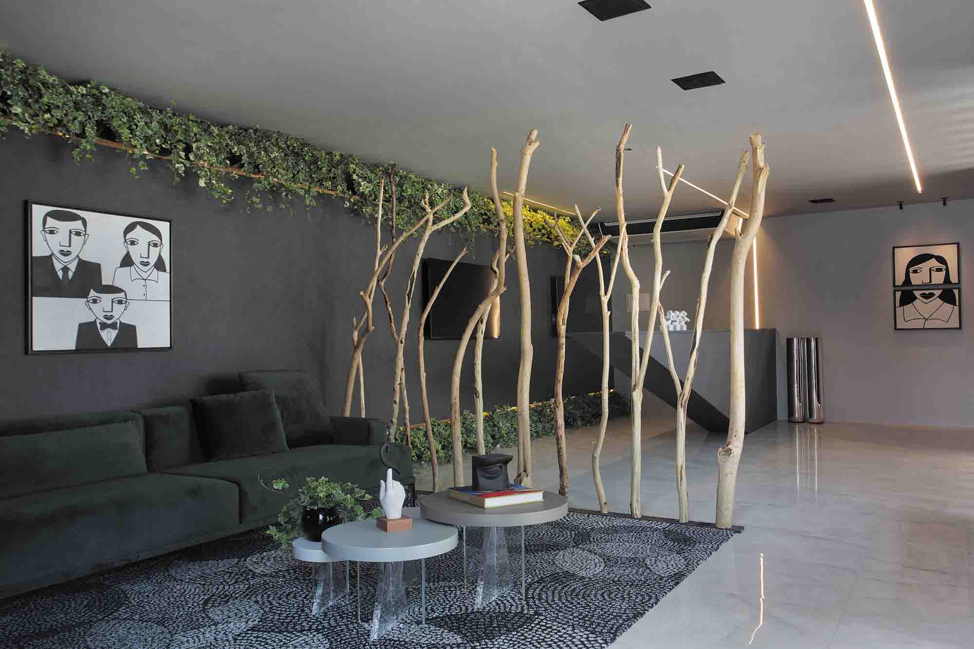 Bilheteria e Lounge de Entrada - DiP Studio Arquitetura. Um encontro entre arte e natureza toma forma logo na entrada da mostra. A curadoria priorizou telas de artistas pernambucanos, que cedem o destaque aos troncos retorcidos in natura com ar de intervenção artística. Um espaço funcional, que encontra em uma paleta cinza próxima ao cimento queimado uma forma de transmitir conforto.