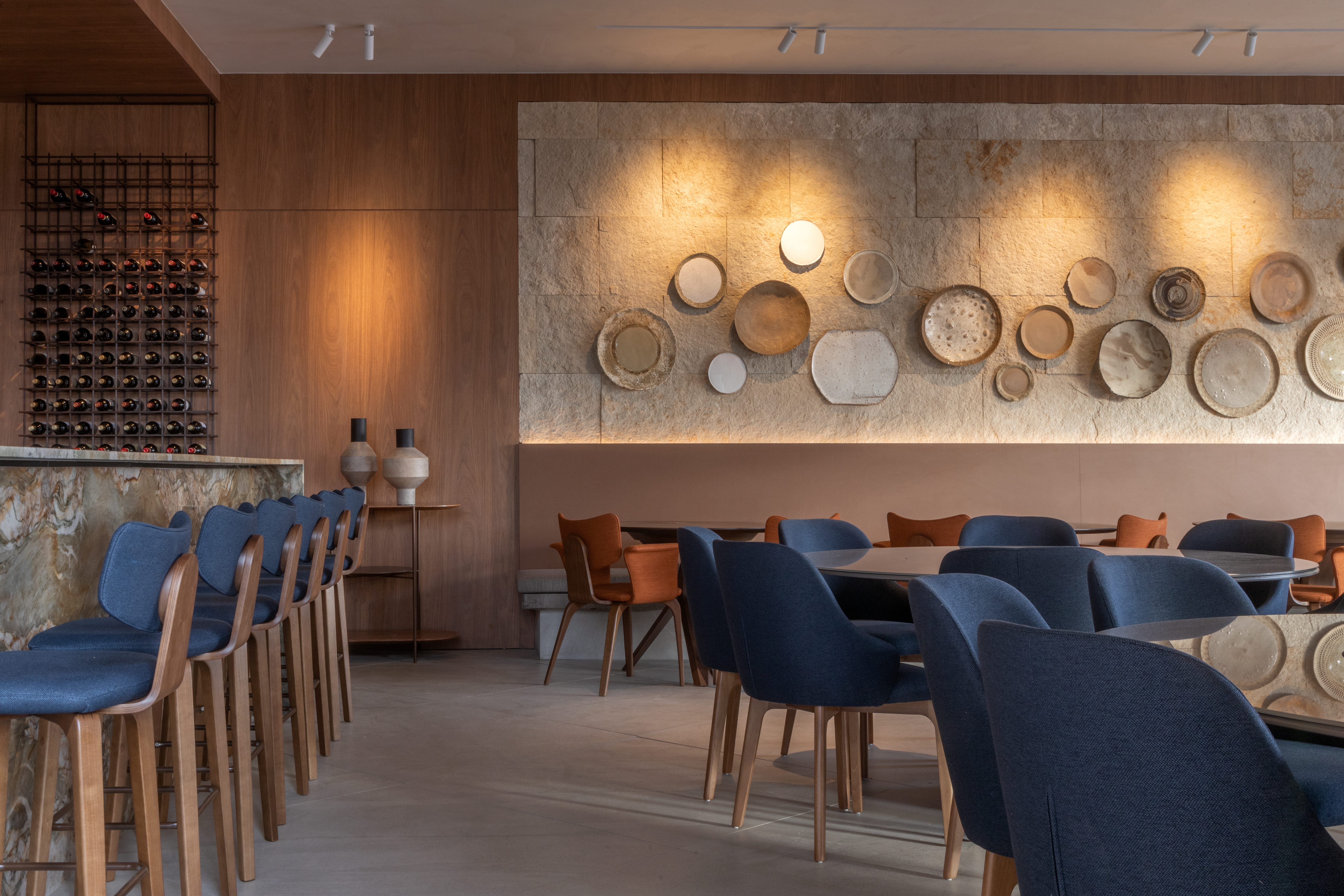 Tatiana Guedes - Restaurante Bar. Um dos lugares que promete reunir visitantes, o local operado pelo Marie Cuisine aposta nas memórias afetivas longevas das porcelanas penduradas nas paredes, com pratos cerâmicos exclusivamente moldados para o ambiente. Entre os elementos utilizados, painéis de mdf jatobá brasileiro, mobília de nogueira, vidro e pedra natural, textura de palha e couro nas cadeiras e linho nas cortinas. No bar, destaca-se o balcão iluminado de quartzito crystal fusion. O verde e o azul integram o local em momentos únicos, com uma vista espetacular do pôr do sol na praça arborizada da mostra.