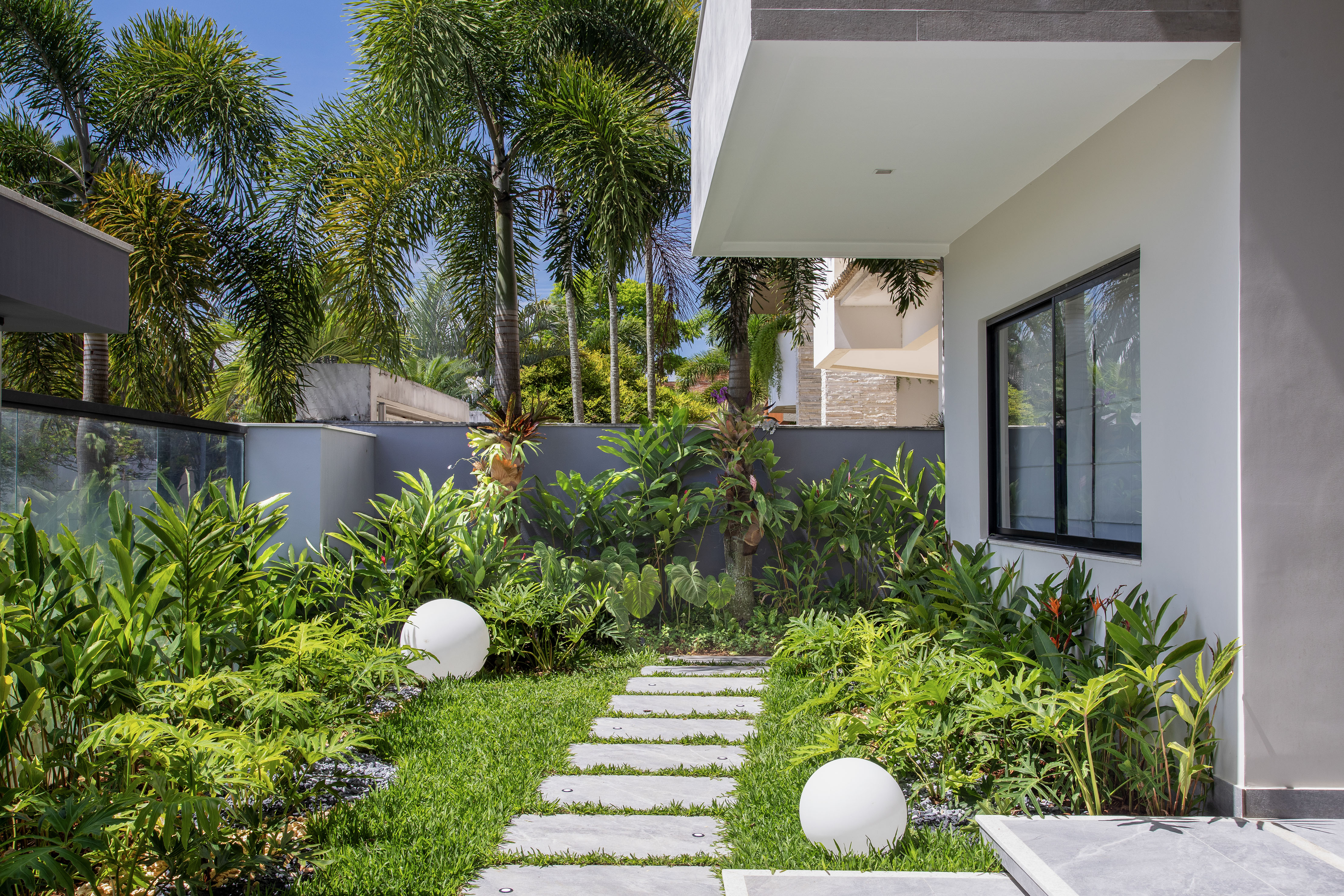 Área de 360 m² ganha jardim com paisagismo tropical e muitas flores. Projeto Maira Duarte. Na foto, jardim com espécies tropicais e luminárias esféricas.